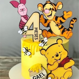Puede incluir: Un pastel con glaseado amarillo decorado con figuras de Winnie the Pooh, Tigger y Piglet. El pastel incluye un adorno de madera con el número 4 y el texto 4 MESES, una figura de tarro de miel con el nombre GAEL y varias abejas pequeñas.