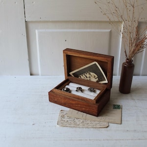 May include: A rectangular wooden jewelry box with a hinged lid, featuring a white velvet ring insert holding three silver rings. Inside the lid, a small black and white photograph is tucked. The box is made of dark brown wood with a visible grain.