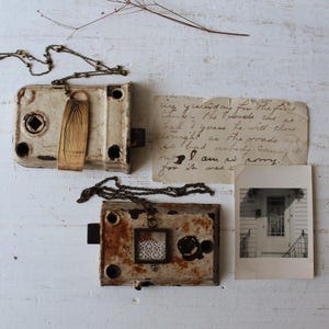 May include: Two antique metal lock boxes with decorative pendants on chains, one with a wing design and the other with a lace insert. A handwritten note and a vintage photograph are also present, all on a white, distressed wooden surface.