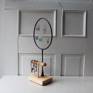 May include: A wooden jewelry stand with a badminton racket for earrings and a wooden rod for bracelets. The stand is made of natural wood and has a rustic look.