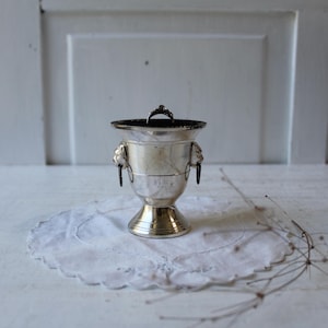 May include: A silver-colored metal vase with a black interior and two lion head handles. The vase sits on a white, scalloped-edge doily. A small, dark ring is inside the vase. The vase is on a white surface.