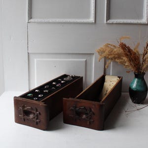 May include: Two wooden jewelry organizers with carved details. One is filled with rings on a black velvet lining. The other is empty, with a cream-colored interior. A vase with dried flowers sits nearby.