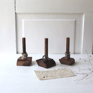 May include: Three wooden ring holders with different rings on each. The holders are on a white surface with a piece of paper and dried flowers.