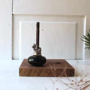 May include: A black glass bottle stopper with a silver floral design and a dark wooden handle. The stopper sits on a square, dark wood base. The background is a white door.