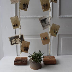 May include: A display stand with earrings on cards and vintage photos. The stand has a wooden base and a white pole with clips. A small potted succulent sits on the white surface.