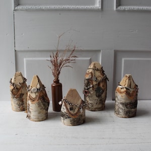 May include: Four cylindrical birch wood jewelry displays with textured bark and light wood tops, each holding a silver chain bracelet. A small brown glass bottle containing dried grass stands between the displays, which are arranged on a white surface.