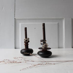 May include: Two rustic ring holders made from dark wood and metal. Each holder features a round base, a vertical post, and a decorative top. Rings of various styles are displayed on the holders, showcasing their design and detail.