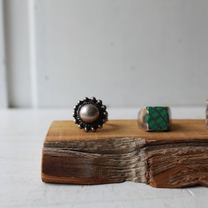 May include: Three silver rings on a wooden display. The ring on the left has a round, silver stone in the center. The ring in the middle has a green stone set in a silver band. The ring on the right is a wide band with a decorative design.