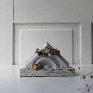 May include: A white wooden triangular jewelry stand with a watch and a bracelet on top. The stand has a distressed white finish and is sitting on a white surface.