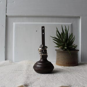 May include: A dark brown, vintage-style ring holder with a decorative base and a vertical post. Several silver rings are displayed on the post. A small potted succulent plant sits to the right, adding a touch of greenery.