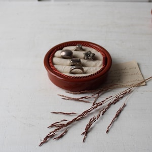 May include: A round, terracotta-colored ring holder with a red rope trim. The interior features a cream-colored fabric with five rings. Dried grass and a handwritten note are next to the ring holder.