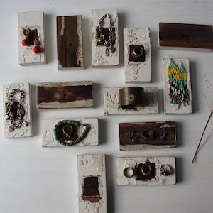 May include: A collection of jewelry displayed on small, rectangular wooden blocks. The jewelry includes earrings, rings, bracelets, and necklaces. The blocks are painted white with distressed edges and feature natural wood accents.