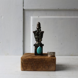 May include: A wooden ring holder with a decorative metal finial topped with turquoise stones and a pendant. The ring holder is on a white surface.