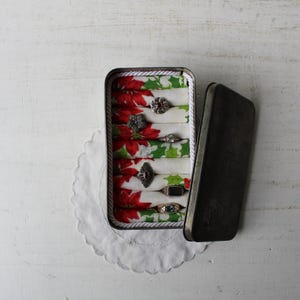 May include: An open, rectangular metal tin with a hinged lid, showcasing several rings nestled on fabric dividers. The fabric features a festive red and green poinsettia pattern. The tin rests on a white, scalloped doily.