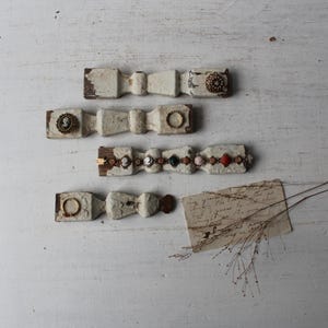 May include: Four decorative, bow-shaped wooden ornaments with antique embellishments. Each bow is painted white with a distressed finish and adorned with vintage jewelry pieces. A handwritten note and dried sprigs are also present.