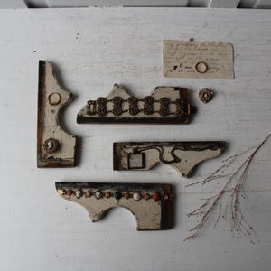 May include: An assortment of vintage, distressed, off-white decorative wooden brackets with ornate details. Some brackets feature metal chain accents, cameo-style embellishments, and colorful stones. A small piece of paper with a ring and a sprig of dried grass are also present.