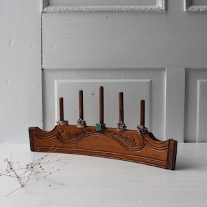 May include: A vintage wooden ring holder with five vertical pegs. The holder is a warm brown color with carved decorative details. Several rings are displayed on the pegs. The background is a white door.