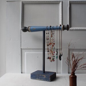 May include: A T-shaped jewelry display stand with a blue horizontal bar and a dark vertical post on a blue rectangular base. Several necklaces with various colorful beads and pendants hang from the horizontal bar, showcasing different lengths.