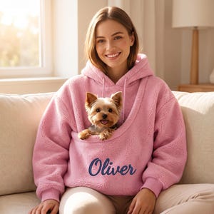 Op de afbeelding: Een lichtroze fleece hoodie met een grote voorzak die is ontworpen om een kleine hond in te dragen. De hoodie heeft lange mouwen, een capuchon met trekkoorden en de naam Oliver geborduurd in donkerblauw schrift op de voorzak.