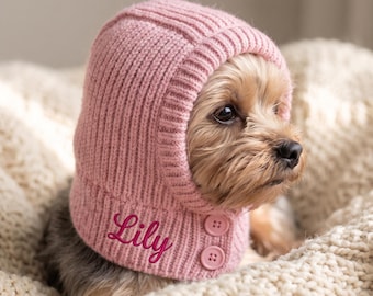 May include: A light pink ribbed knit pet bonnet featuring the name Lily embroidered in dark pink script. The bonnet has a rounded hood design with two matching pink buttons on the side, framing the face of a small brown and tan dog.