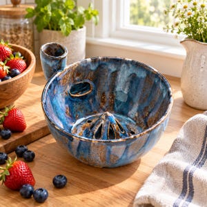 Può includere: Uno spremiagrumi in ceramica con smalto reattivo blu profondo e marrone. La ciotola presenta un cono centrale con fori di drenaggio, un manico laterale e un'apertura ovale sul lato. La finitura è lucida con visibili variazioni di colore.