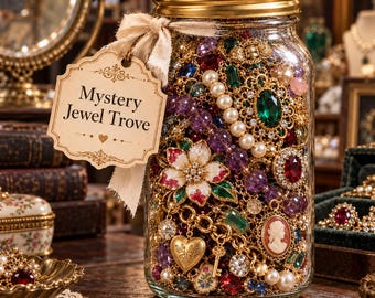 Puede incluir: Un frasco de vidrio transparente con tapa dorada lleno de bisutería variada, incluyendo perlas, gemas de colores, cadenas doradas y un camafeo. Una etiqueta de papel beige atada con una cinta dice Mystery Jewel Trove.