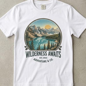 Könnte beinhalten: Ein weißes Kurzarm-T-Shirt mit einer kreisförmigen Grafik, die eine Bergseelandschaft mit Tannen und einer Sonne zeigt. Unter der Grafik steht der Text "WILDERNESS AWAITS EST. 2023 ADVENTURE & CO." in dunkelblauer Schrift auf der Brust.