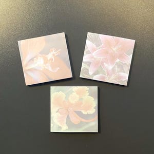 May include: Three square sticky notes with floral designs. Each note features a different close-up of a pink and peach flower. The notes are arranged on a dark gray surface.