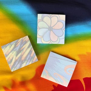 May include: Three square notepads with colorful designs. One features a flower pattern, another has diagonal lines, and the third has a wave-like design. The background is a vibrant rainbow gradient.