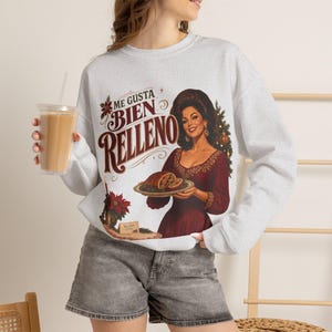 May include: A light gray crewneck sweatshirt featuring a graphic print of a person in a dark red dress holding a platter with a roasted dish. The text reads ME GUSTA BIEN RELLENO. The design includes a Christmas tree, poinsettia, and decorative flourishes.