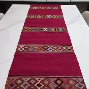May include: A long, rectangular table runner in a deep burgundy red color. The runner features three distinct horizontal bands of intricate, multi-colored geometric diamond patterns in shades of orange, yellow, white, and black, finished with fringe.