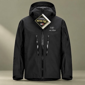 May include: A black hooded jacket featuring a full-length front zip, two vertical chest pockets with black zips, and a small pocket on the left sleeve. The Arc'teryx logo is printed on the chest, and a Gore-Tex tag is attached to the collar.