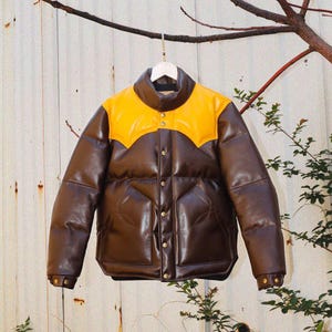 May include: A brown puffer jacket featuring a contrasting bright yellow yoke across the shoulders and chest. The jacket has a high collar, a front button closure with metallic snaps, long sleeves with snap cuffs, and two large front pockets.