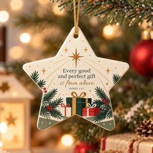 Peut inclure: Une décoration en céramique en forme d'étoile avec une ficelle dorée. Le motif montre des branches de pin vertes, des baies rouges et trois cadeaux. Le texte indique : Every good and perfect gift is from above. JAMES 1:17, avec une étoile dorée.