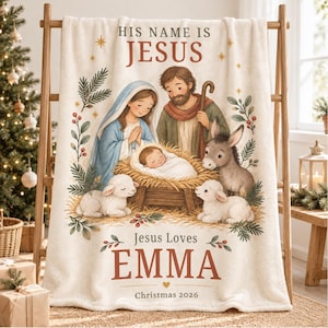 May include: A cream-coloured fleece blanket featuring a nativity scene with Mary, Joseph, and baby Jesus in a manger. The text reads HIS NAME IS JESUS, Jesus Loves EMMA, and Christmas 2026. Two lambs and a donkey are illustrated near the manger.