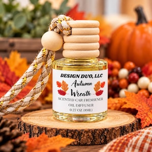 Könnte beinhalten: Eine durchsichtige Glasflasche für Auto-Dufterfrischer mit hellem Holzdeckel und geflochtener Kordel. Das weiße Etikett trägt den Text DESIGN DUO, LLC Autumn Wreath SCENTED CAR FRESHENER OIL DIFFUSER 8ML mit orangefarbenen und roten Blattgrafiken.