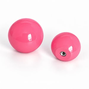 May include: Two spherical knobs with a glossy hot pink finish. One knob is positioned to show its smooth, rounded surface, while the other is angled to reveal a circular flat base with a centered, threaded metal insert for mounting purposes.