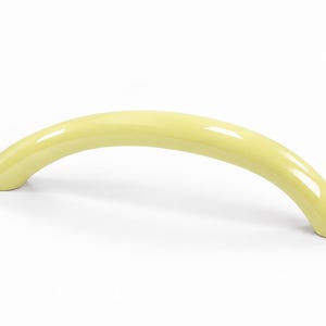 Puede incluir: Un tirador de mueble arqueado de color amarillo pálido con un acabado liso y brillante. El herraje presenta un diseño curvo en forma de semicírculo que se estrecha en ambos extremos, ofreciendo un agarre sencillo para puertas o cajones.