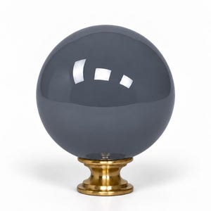 May include: A spherical dark gray glass knob with a smooth, reflective surface. The glass sphere is securely attached to a circular, metallic gold-toned base that features a stepped, pedestal-like design with a flat bottom for mounting.