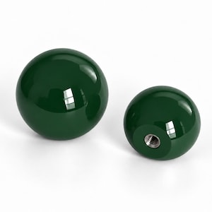May include: Two spherical knobs with a glossy dark forest green finish. One knob is positioned to show its smooth, rounded surface, while the other is oriented to reveal a flat base featuring a centered, circular metal threaded insert.