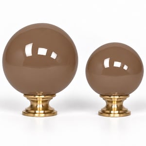 May include: Two spherical finials featuring smooth, glossy, taupe-colored glass orbs mounted on polished brass bases. The larger orb sits on the left, while the smaller orb is positioned to the right, both displaying reflective surfaces.