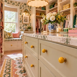 May include: A sage green kitchen island with a white marble countertop and bright yellow spherical knobs. The island features multiple drawers and is situated in a walk-in closet with white shelving units, floral wallpaper, and a window seat.