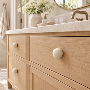 May include: A light oak wood bathroom vanity cabinet featuring three drawers and one door, each equipped with a smooth, round, cream-colored ceramic knob. The vanity is topped with a light beige stone countertop and a brass faucet fixture.