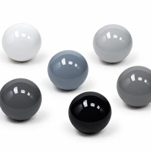 May include: A collection of six smooth, spherical objects arranged in a cluster. The spheres feature a glossy finish and vary in color, ranging from solid white to light gray, medium gray, dark gray, and a single deep black sphere at the center.