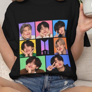 May include: A black t-shirt featuring a grid of seven individual portraits of the members of the music group BTS, with the purple and white BTS logo centered in the middle square. The graphic is arranged in a three-by-three layout with colorful backgrounds.