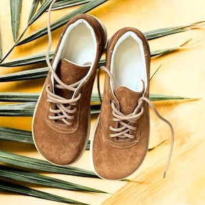 May include: A pair of brown suede sneakers with a wide, rounded toe box, white interior lining, and matching brown laces. The shoes feature a low-top design with visible white stitching along the edges and a flat, white rubber sole base.