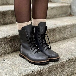 May include: A pair of dark gray mid-calf lace-up boots featuring black laces, a rounded toe cap, and a thick black lug sole. The boots are worn with beige ribbed socks and sheer black tights, showcasing a clean design with visible stitching.