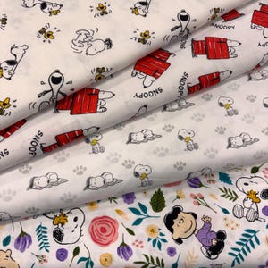 以下が含まれることがあります： スヌーピー、ウッドストック、赤い犬小屋と「SNOOPY」の文字が繰り返される白い生地。別のセクションには、灰色の足跡と共に眠るスヌーピーが描かれています。3つ目のセクションには、スヌーピー、ウッドストック、ルーシーと花々が描かれています。