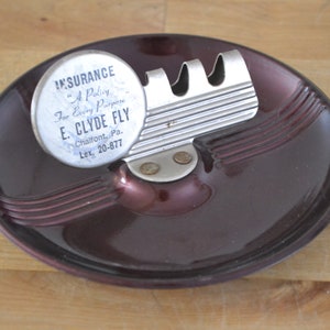 May include: A burgundy ceramic ashtray with a silver metal cigarette holder. The ashtray has a raised design around the edge. The cigarette holder has three slots for cigarettes. A round, white sticker with black text is attached to the cigarette holder. The text reads "INSURANCE "A Policy" For Every Purpose E. CLYDE FLY Chalfont, Pa. Lex, 20-877"