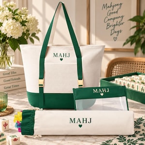 May include: A white and dark green canvas tote bag, a matching rectangular pouch with a clear front panel, and a long cylindrical drawstring bag. Each item features the text MAHJ with a small heart icon in dark green embroidery.